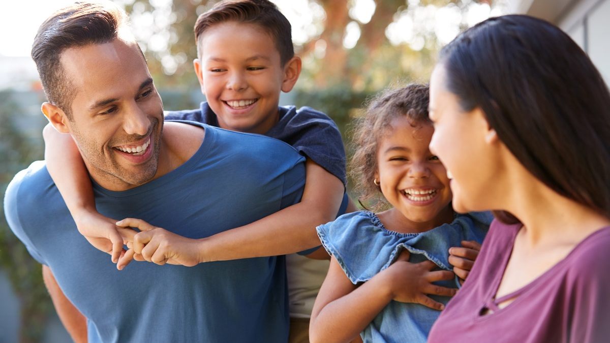 Smiling Hispanic Family With Parents Giving Children Piggyback Rides In Garden At Home